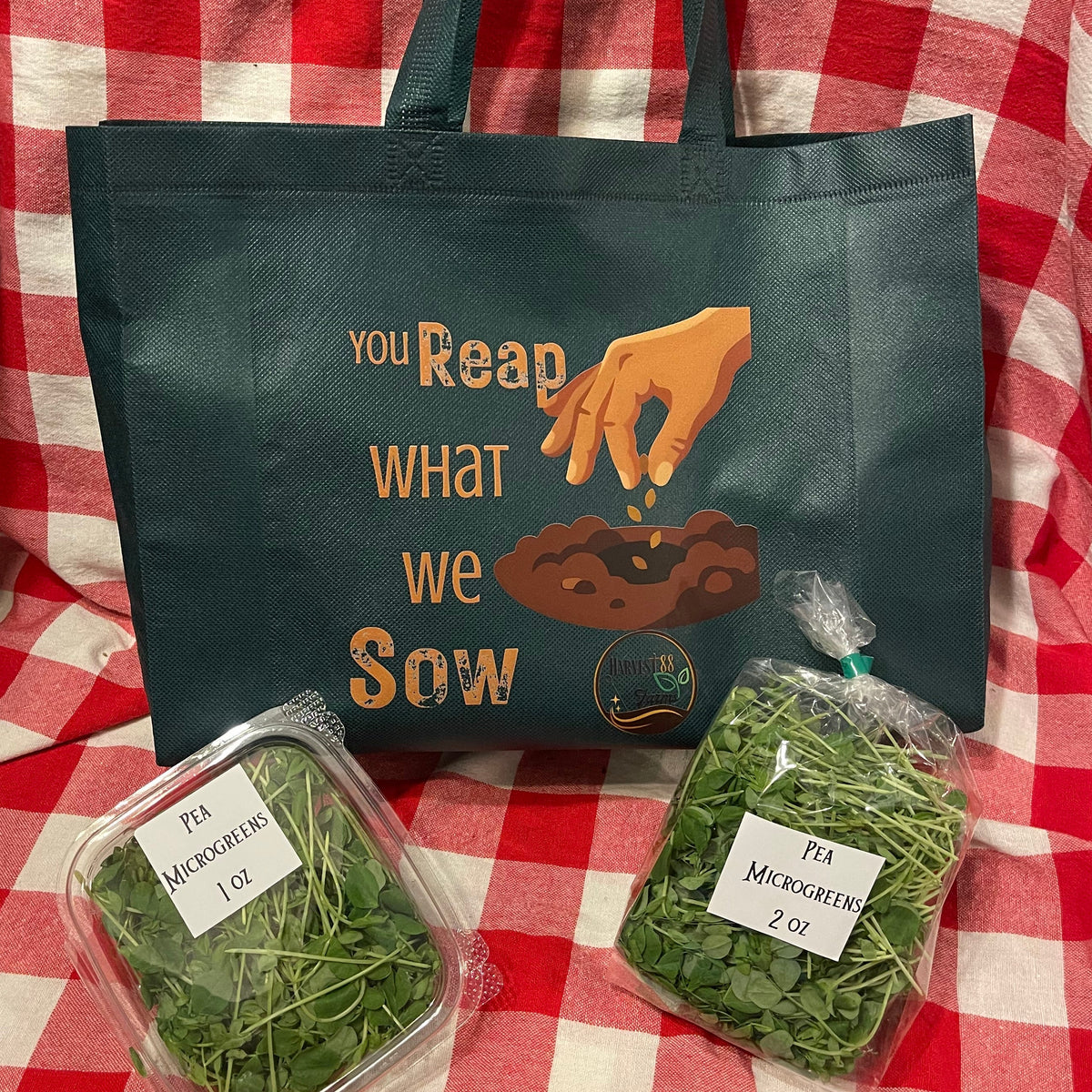 A dark green reusable tote bag with a printed graphic and quote sits on a red and white checkered blanket, with two clear plastic bags of fresh green herbs placed in front.