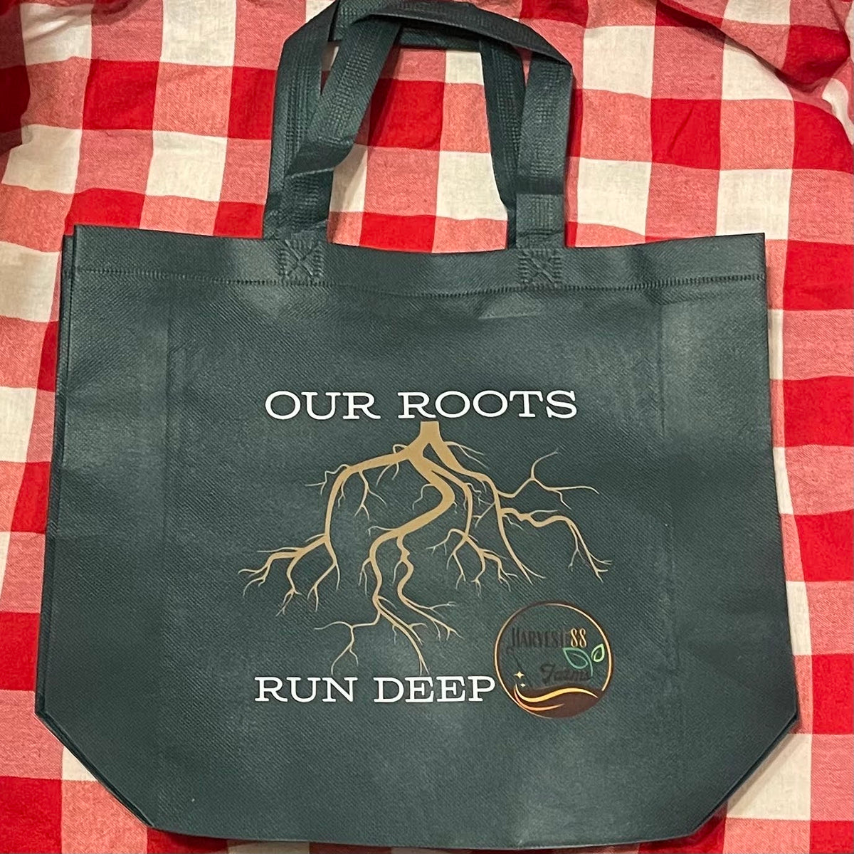A dark Green reusable tote bag with 'OUR ROOTS RUN DEEP' and a tree root graphic printed on the front, laid flat on a red and white checkered blanket.