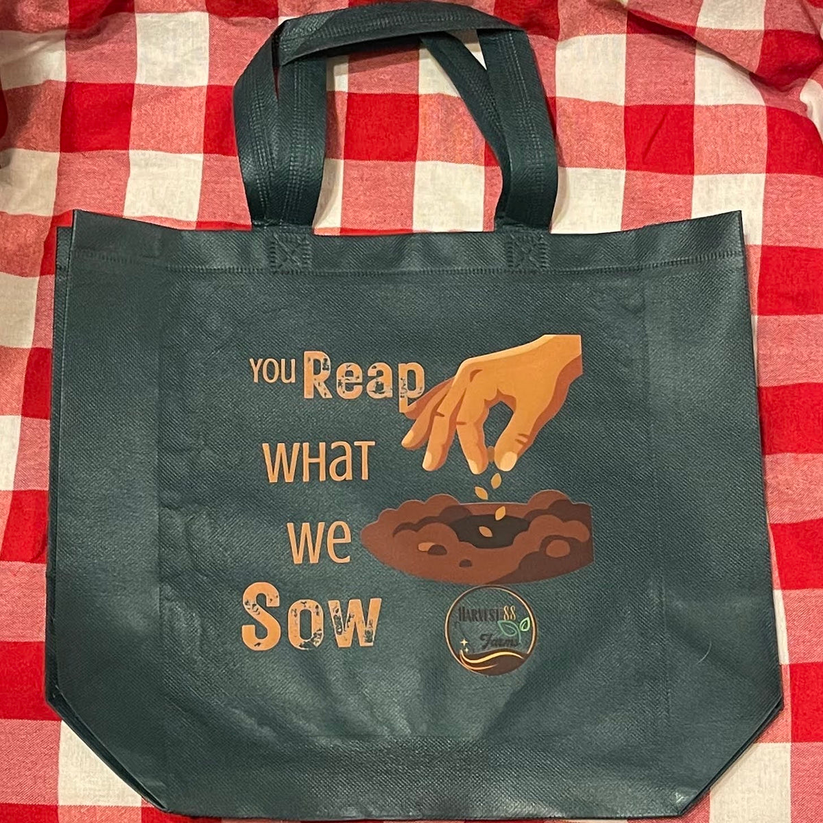 A dark green reusable tote bag with black handles, featuring a graphic of a hand sowing seeds and the text 'you Reap what we Sow'. The bag is laid flat on a red and white checkered blanket.