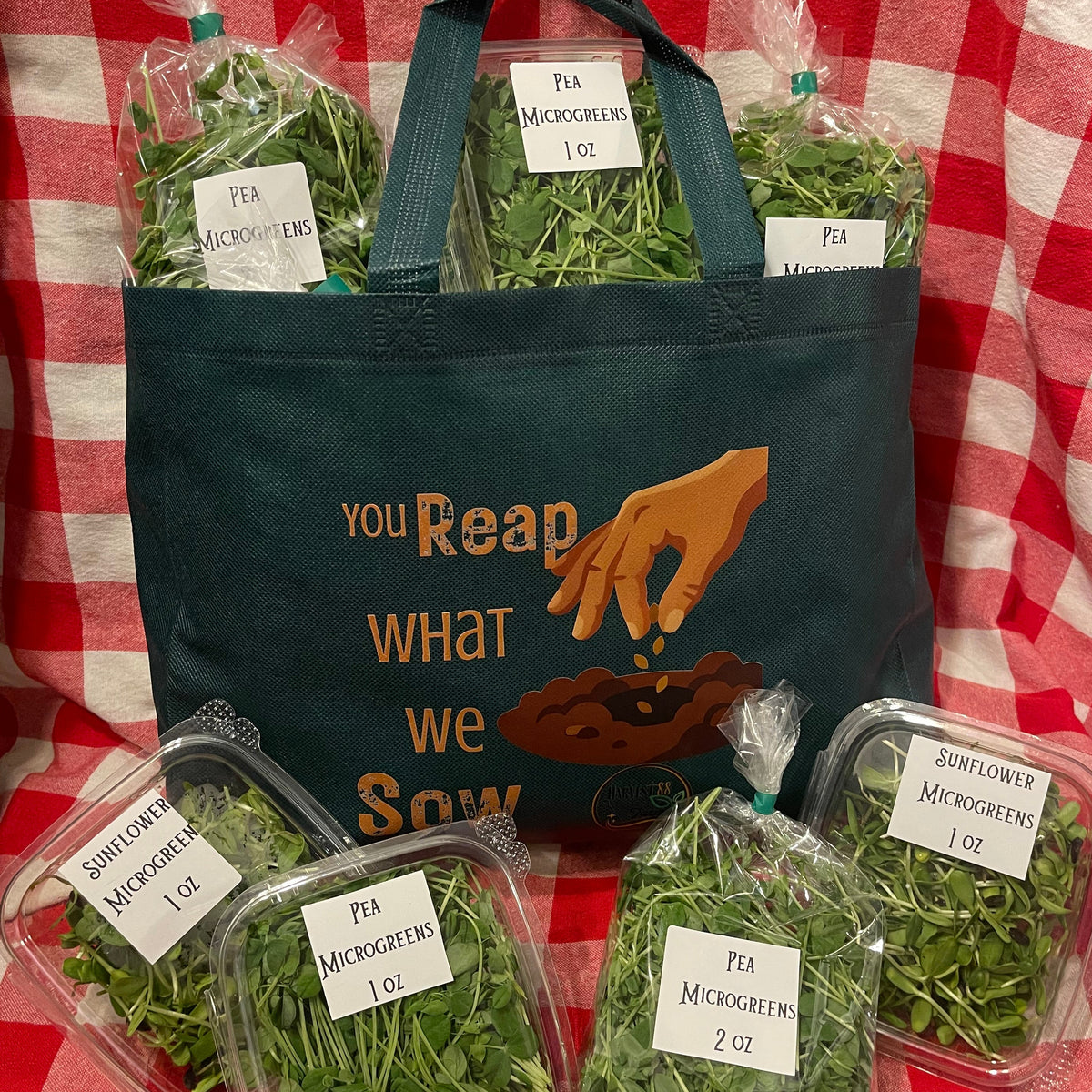 A Green reusable tote bag with the printed phrase 'YOU Reap WHAT we Sow' and a hand planting seeds, surrounded by several clear plastic bags and containers filled with green microgreens on a red-and-white checkered fabric backdrop.