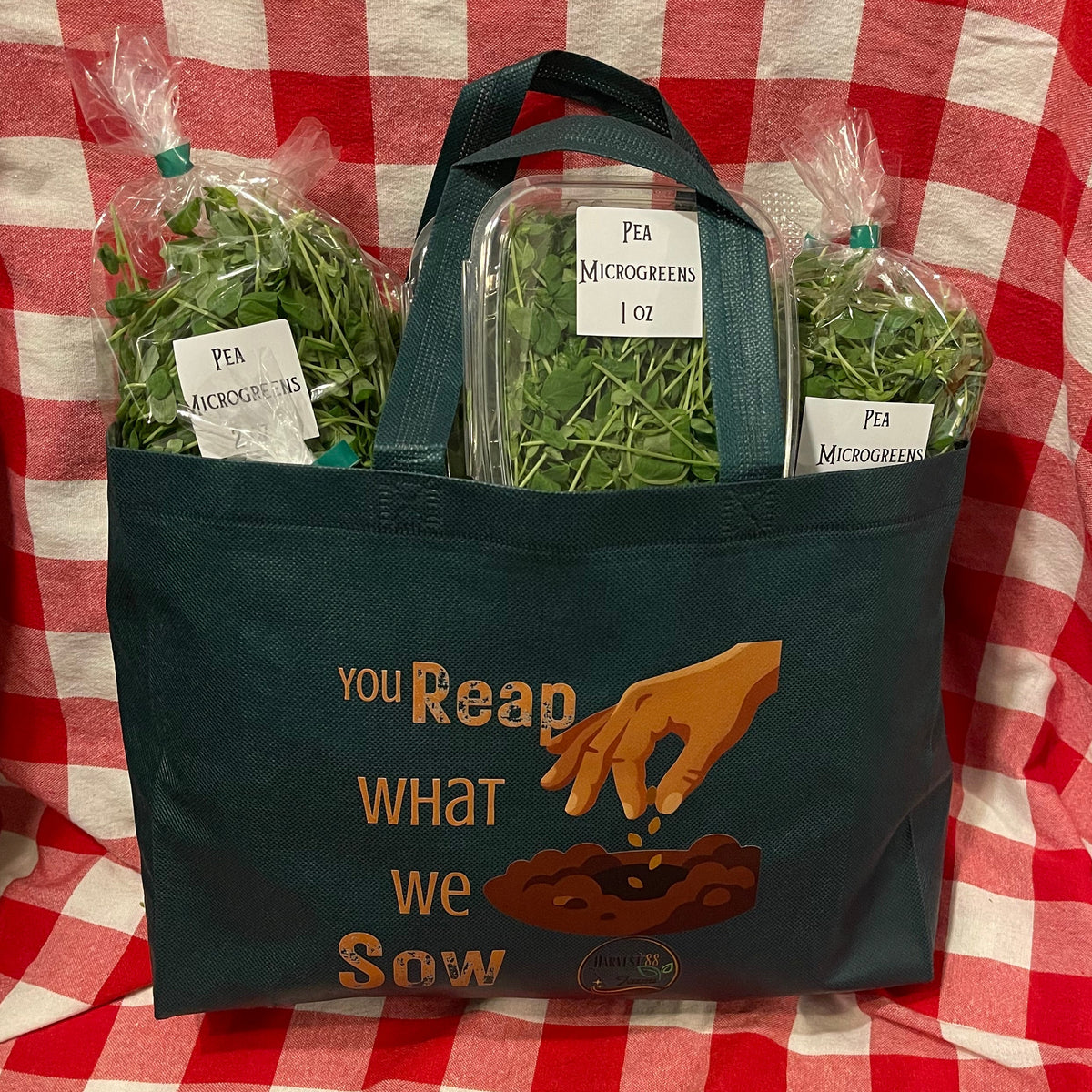 A dark green canvas tote bag with a printed graphic and quote sits on a red and white checkered blanket. The bag is filled with three packages of microgreens, including two labeled 'Pea Microgreens' and one in a clear container.