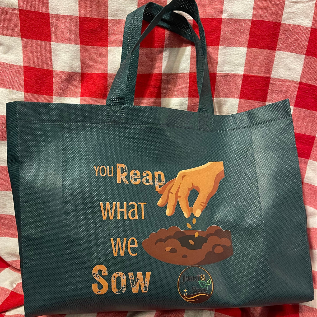 A dark green reusable tote bag with a printed graphic of a hand sowing seeds over the words 'YOU REAP WHAT WE SOW' in orange and brown, displayed on a red and white checkered blanket.