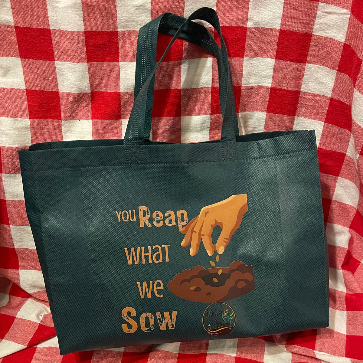 A dark green reusable tote bag with a printed graphic of a hand sowing seeds and the text 'YOU Reap WHAT we SOW', displayed on a red and white checkered blanket.