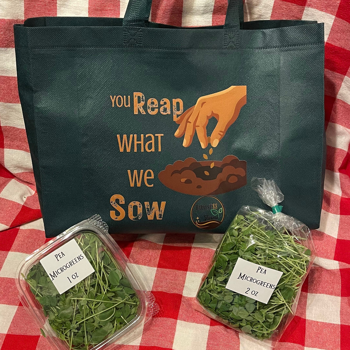 A dark green reusable tote bag with a gardening graphic and text sits on a red and white checkered blanket, alongside two clear plastic bags of fresh green micro greens.